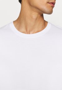 Närbild av en persons nacke och käklinje som bär en vanlig vit T-shirt med rund hals, riktad något åt höger.