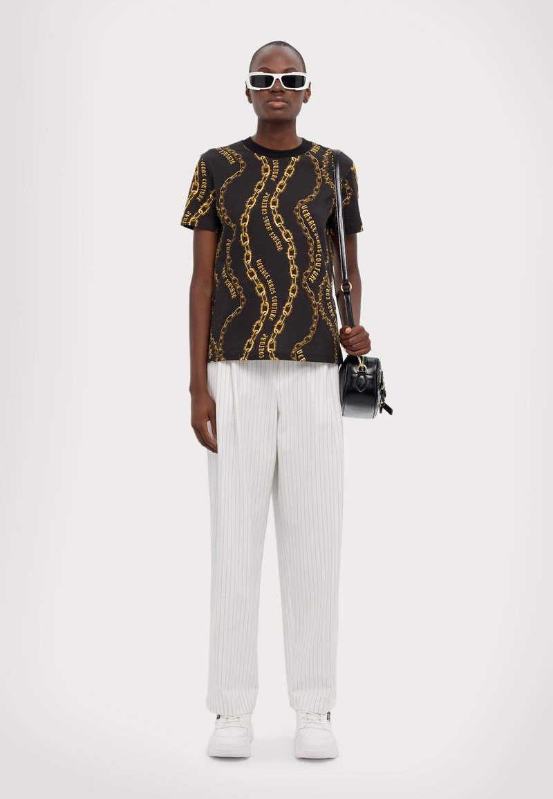 Persona con una camiseta negra de patrón de cadenas, pantalones blancos de rayas, zapatillas blancas, gafas de sol blancas y llevando un bolso negro al hombro.