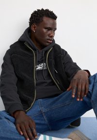 Gilet en polaire noir avec des finitions olive sur un sweat à capuche noir, associé à un jean bleu. Assis sur une surface bleu clair, portant des chaussures beige.