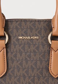 Καφέ τσάντα χειρός με παδιά σχέδια από υφή συνθετικού υλικού, με χρυσαφένια μεταλλικά στοιχεία και λογότυπο. Τακάκια σε ταμπά χρώμα στα χερούλια.