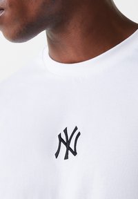 Valge puuvillane T-särk, millel on vasakul rinnal väike must broderitud NY logo. Ümar kaelus ja sile tekstuur.