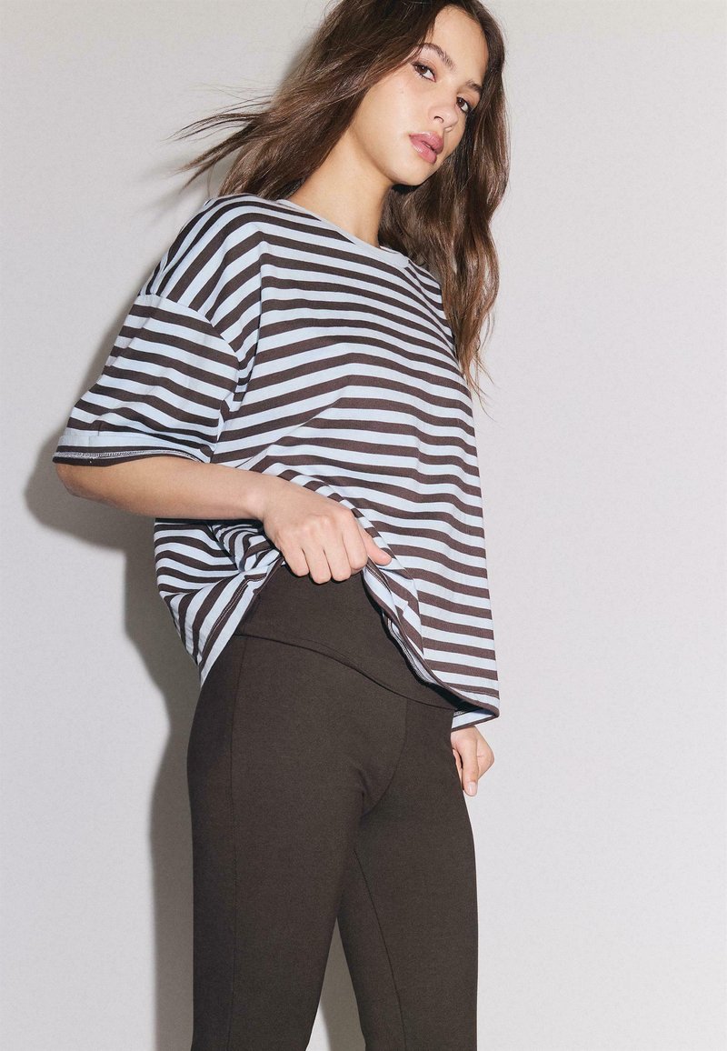 Jeune femme aux longs cheveux bruns portant une chemise rayée bleue et marron et un pantalon marron foncé ajusté, sur un fond clair uni.