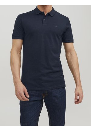 Man wearing a plain dark navy blue polo shirt and dark blue jeans standing against a light gray background.
