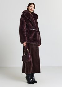 Giacca in pelliccia ecologica bordeaux con colletto largo e cintura, abbinata a una lunga gonna di velluto marrone e stivali neri con tacco alto, con una borsa in mano.