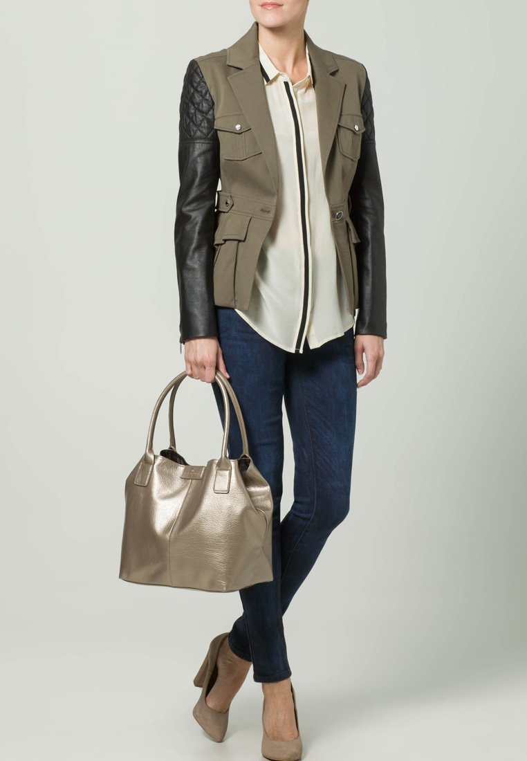 Donna che tiene in mano una borsa metallica dorata, indossando un giacca olivastro con maniche in pelle nera, blusa crema, jeans scuri e tacchi beige.