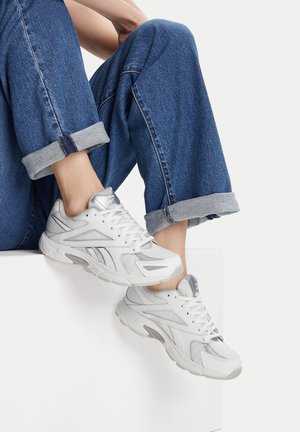 Białe sportowe trampki wykonane z siateczki i materiałów syntetycznych, z jasnoszarymi akcentami. Gruba podeszwa i sznurowane zapięcie są wyraźnie widoczne.