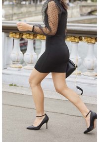 Black mini dress with polka dot sheer sleeves, paired with black stiletto heels and a small black handbag with a chain strap.