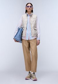 Gilet imbottito beige su una camicia a righe celesti, abbinato a pantaloni beige e sneakers beige. Gli accessori includono occhiali da sole viola e una borsa blu.