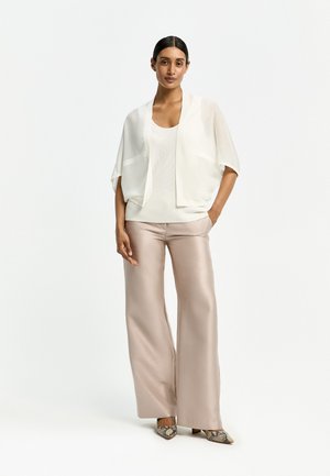 Frau trägt eine weiße, locker sitzende Strickjacke mit kurzen Ärmeln, ein weißes Tanktop, beige Hosen mit weitem Bein und gemusterte spitze Schuhe, steht mit einer Hand in der Tasche.