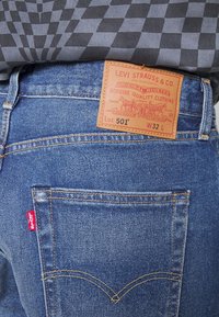 Jean en denim bleu avec une étiquette en cuir portant l'inscription "Levi Strauss & Co. Lot 501 W32 L" et des surpiqûres contrastantes jaunes. Onglet rouge sur la poche.