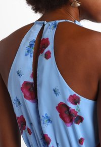 Mörkhårig person iklädd en ärmlös ljusblå klänning med röda och blå blommönster, som visar baksidan med en knapplåst nyckelhålsringning.