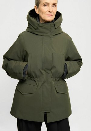 Persona madura con chaqueta de invierno verde con capucha y manos en los bolsillos, de pie frente a un fondo liso.