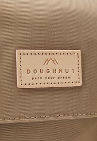 Mochila de tela beige con una superficie texturizada; cuenta con un parche de cuero rectangular etiquetado "DOUGHNUT" con un logo de montaña y costuras.