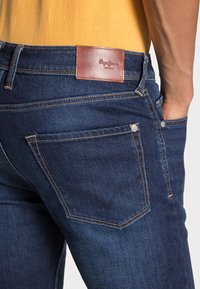 Jeans de denim azul escuro com um remendo de couro castanho na cintura e costuras contrastantes. Inclui bolsos traseiros com detalhes em metal.