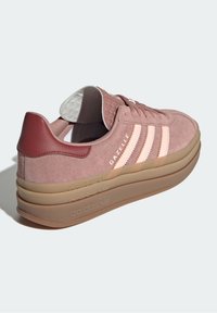 Baskets Adidas Gazelle en daim rose avec un talon bordeaux contrastant, des rayures blanches et une semelle épaisse et translucide. Texture douce et forme arrondie.