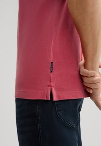Różowy bawełniany T-shirt z teksturowanym materiałem, bocznymi rozporkami u dołu i małą czarną metką na dolnym szwie, w połączeniu z ciemnymi dżinsami.