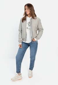 Lys grå bomberjakke med ribbede manchetter, båret over en hvid hoodie med blomsterprint. Kombineret med blå denimjeans og beige sneakers.