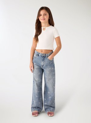 Dziewczyna o długich brązowych włosach, ubrana w biały krótki top z krótkim rękawem, szerokie jeansy z wzorem kokardek i błyszczące różowe buty, stojąca na białym tle.