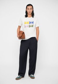 Vyras vilkintis baltą marškinėlį su spalvotu kasetės spaudiniu, juodomis smulkiai dryžuotomis plačiomis kelnėmis, turkio spalvos sportiniais bateliais, laiko rudą odinę rankinę.