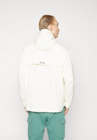 Nike jakke i blødt cremet stof, med hætte, rygventilation og logodetalje. Løs pasform med elastiske maofer og kant.