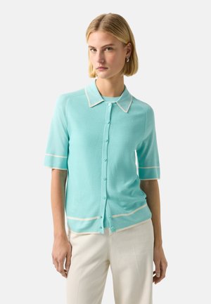 Kobieta w jasnoniebieskim kardiganie na guziki z krótkim rękawem i białym obszyciem oraz białych spodniach, stojąca na tle jednolitego jasnego tła.