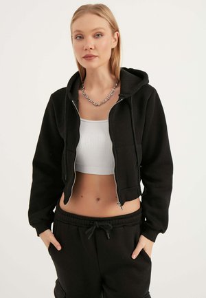 Felpa nera corta con zip, abbinata a una canotta grigia a costine. La felpa presenta un cappuccio con coulisse e una texture liscia.