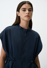 Robe bleu marine à manches courtes avec un col montant, une fermeture éclair sur le devant et une taille cintrée. Fabriquée en tissu lisse avec des détails de coutures visibles.
