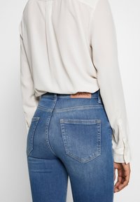 Vaqueros de denim azul claro con un corte ajustado, que cuentan con dos bolsillos traseros y una etiqueta de cuero color madera en la cinturilla. Blusa blanca de manga larga visible.