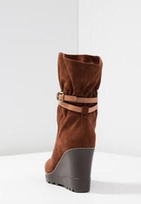 Bruna mockaskinns klackstövlar med en mjukt nedtill design, utrustade med ett justerbart tanfärgat band och en rundad tå. Klackens topp är mörk och texturerad.