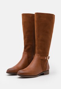 Bottes marron hautes en daim et cuir, dotées d'une fermeture éclair latérale, d'un talon en bois plat et d'une sangle décorative avec un accent métallique.