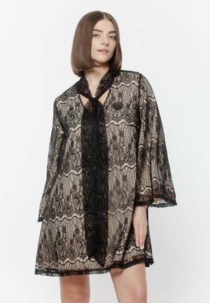 Kobieta o prostych brązowych włosach ubrana w przezroczystą czarną koronkową sukienkę z dołączonym szalikiem, stojąca na tle jednolitego białego tła.