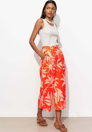 Femme portant un haut blanc sans manches, un pantalon large orange vif avec des motifs floraux clairs, des sandales marron à enfiler et des colliers blancs.