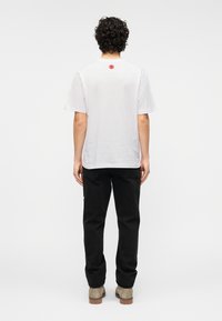 Λευκό βαμβακερό T-shirt με στρογγυλή λαιμόκοψη, κοντά μανίκια και κόκκινο κυκλικό σχέδιο στην πλάτη, σε συνδυασμό με μαύρο παντελόνι και μπεζ παπούτσια.