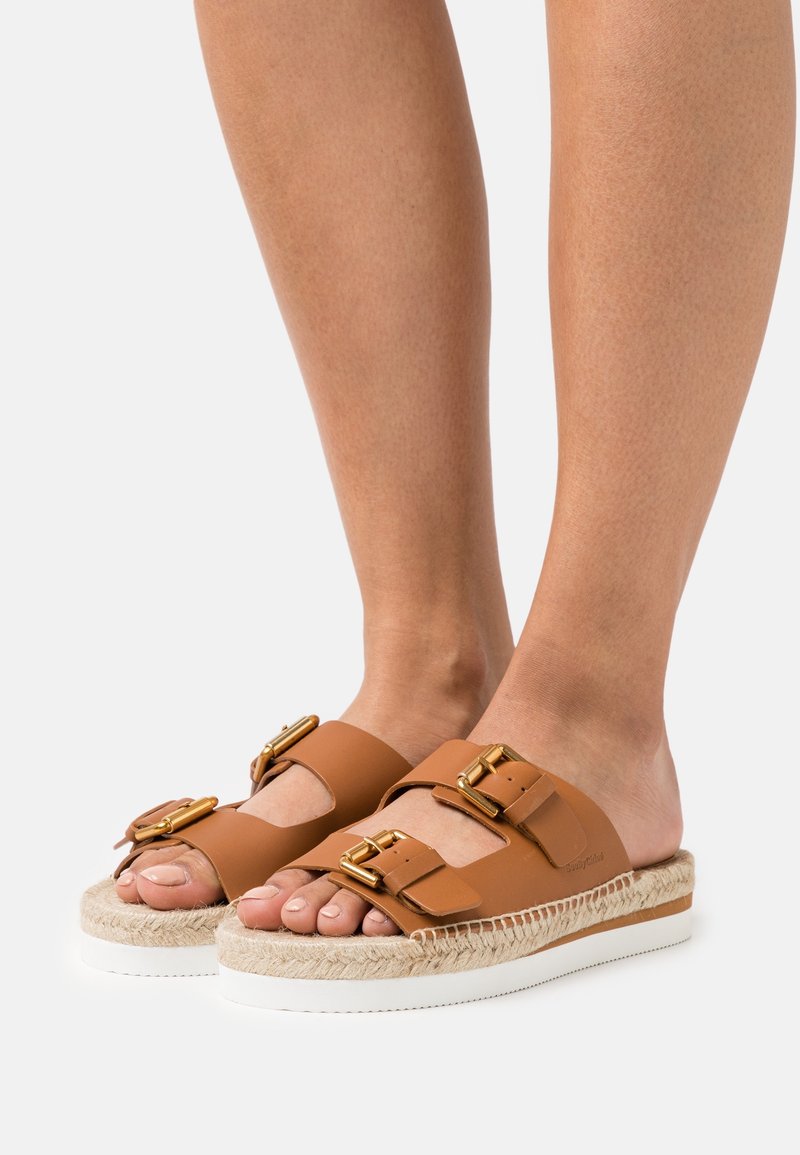 Pieds portant des sandales en cuir beige à enfiler avec deux boucles dorées réglables et semelles compensées bordées de corde sur fond blanc.