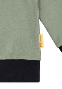 Felpa verde in cotone con polsini e orlo a coste neri. Presenta un'etichetta gialla sul lato con testo rosso. Texture morbida e liscia.