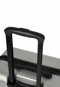 Maleta plateada con una superficie texturizada, mango telescópico negro y botón de liberación. Se pueden ver el cierre de cremallera y las esquinas reforzadas.