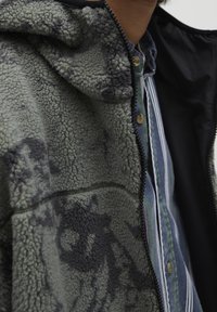 Polarowa kurtka w oliwkowym zieleni z czarnym abstrakcyjnym wzorem, z kapturem, zapięciem na zamek i teksturowaną powierzchnią. Widoczna koszulka pod spodem.