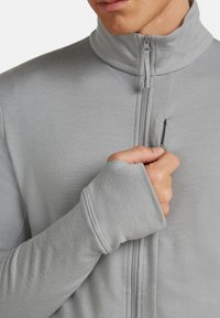 Persona che indossa una giacca leggera grigia con zip e foro per il pollice nella manica, mentre sta parzialmente aprendo la cerniera di una piccola tasca sul petto.