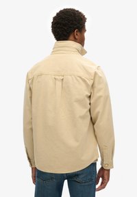 Chaqueta de algodón beige con un cuello alto, yugo trasero y puños abotonados. Presenta un diseño limpio y sencillo con una textura suave.