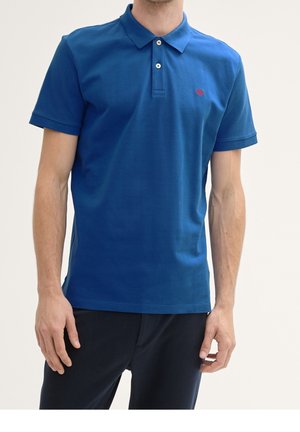 Homme portant un polo bleu à manches courtes avec deux boutons blancs et un petit logo rouge sur la poitrine gauche, assorti à un pantalon foncé.