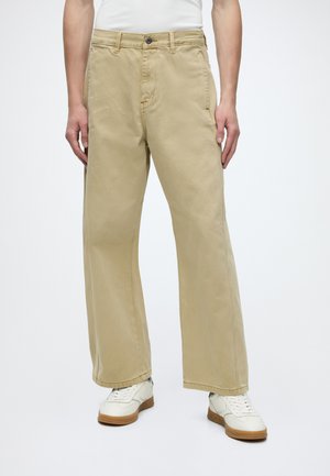 Persona che indossa pantaloni beige larghi e sneaker bianche con suole marroni, in piedi su uno sfondo bianco uniforme.