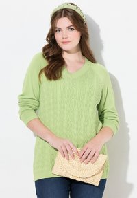 Vaaleanvihreä neulepusero, jossa on V-pääntie, palmikkoneule ja pitkät hihat. Malli pitää beigen väristä kudottua clutch-laukkua. Siniset farkut näkyvät.