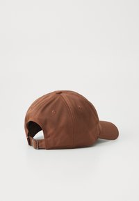 Brun bomull baseball caps med justerbar metallspenne ligger på en hvit overflate med fronten mot venstre.