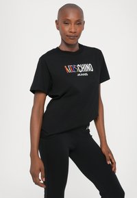 Juodas medvilninis marškinėlis trumpomis rankovėmis, su daugiaspalviu "KATE CHINO JEANS" logotipu krūtinės srityje. Paprastas, laisvas kirpimas.