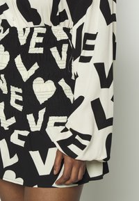 Abito bianco e nero con grandi lettere in grassetto e forme a cuore, caratterizzato da una vita arricciata e lunghe maniche a sbuffo, posato su una mano di carnagione scura vicino alla coscia.