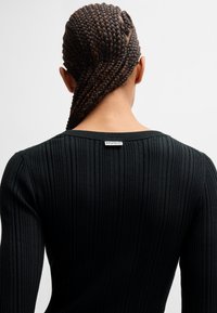 Black ribbed long-sleeve top with a wide neckline. Features a small white label at the back with "HUGO" text. Smooth texture.