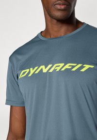 Harmaa urheilupaita pyöreällä kaula-aukolla, jossa on rohkea keltainen "DYNAFIT" logo rinnassa. Pehmeä, sileä kangas.