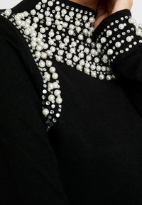 Schwarzer Pullover mit hohem Kragen, verziert mit Clustern aus weißen Kunstperlen und klaren Strasssteinen entlang der Schultern.