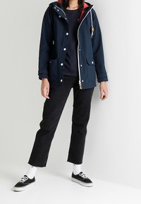 Navy blue waterproof jacket with a red interior, front zip closure, and snap buttons. Features slanted pockets and a hood. Worn with black jeans.