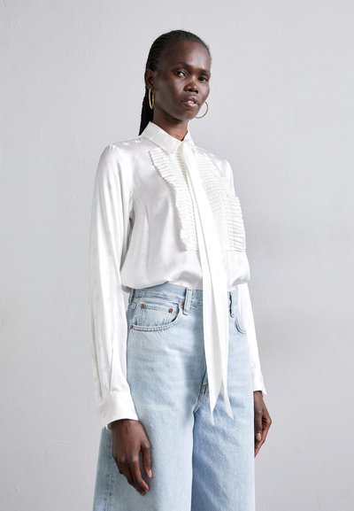 Blouse en satin blanc avec détail froncé sur le devant, manches longues et col noué. Associée à un jean taille haute bleu clair.